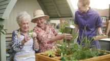 Two older people enjoying planting and watering seedlings and flowers in a raised planter as a member of staff joins the fun