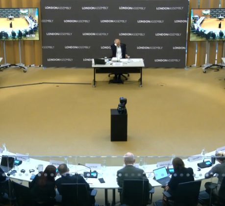 A man at a desk facing questioners sitting in a horseshoe